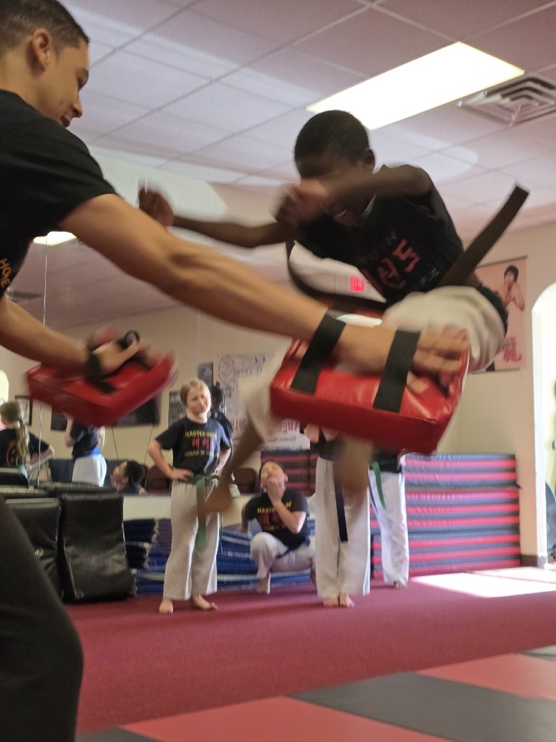 A young student launching a flying kick into a held pad.