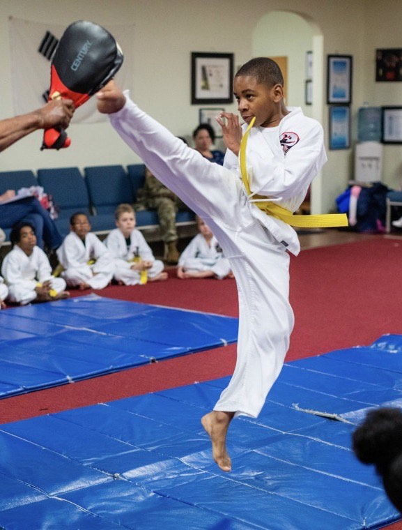A young student throwing a high kick into a pad held by Master Irvin, with the rest of the class watching from the floor.